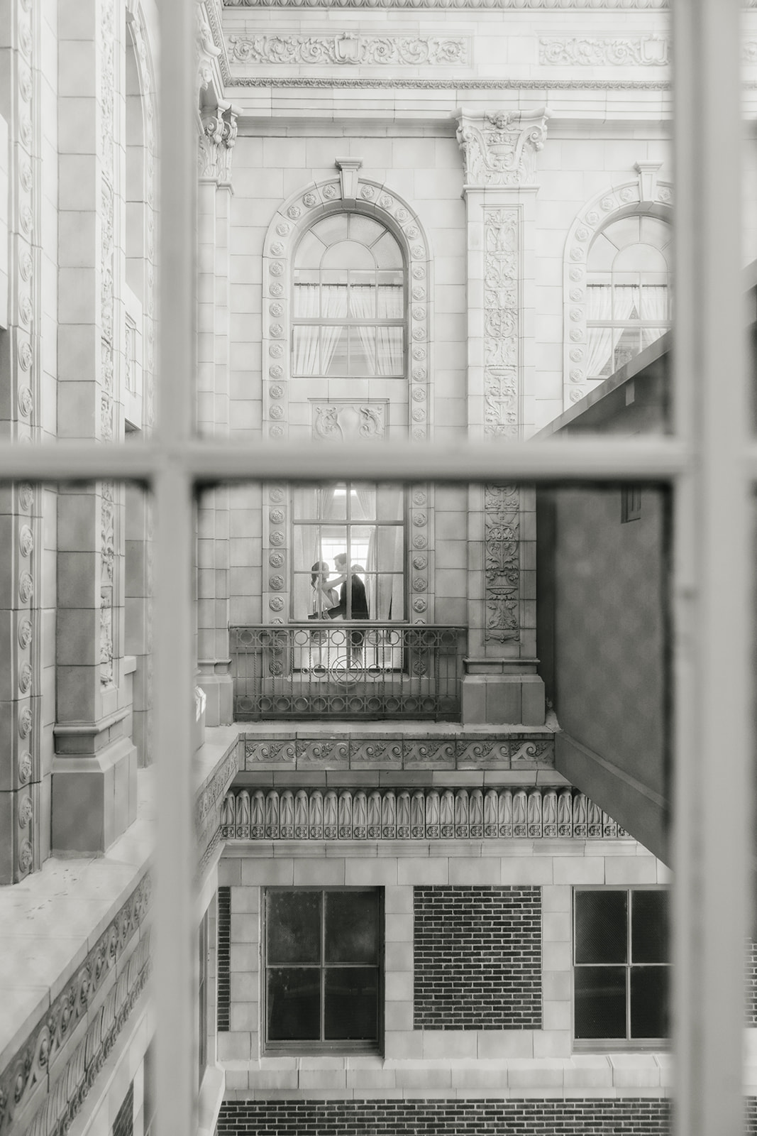 Two people stand facing each other on a balcony inside a historic building, viewed through a window from across the way.