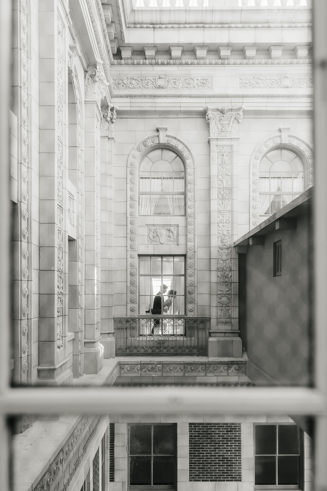 Two people stand facing each other on a balcony inside a historic building, viewed through a window from across the way.