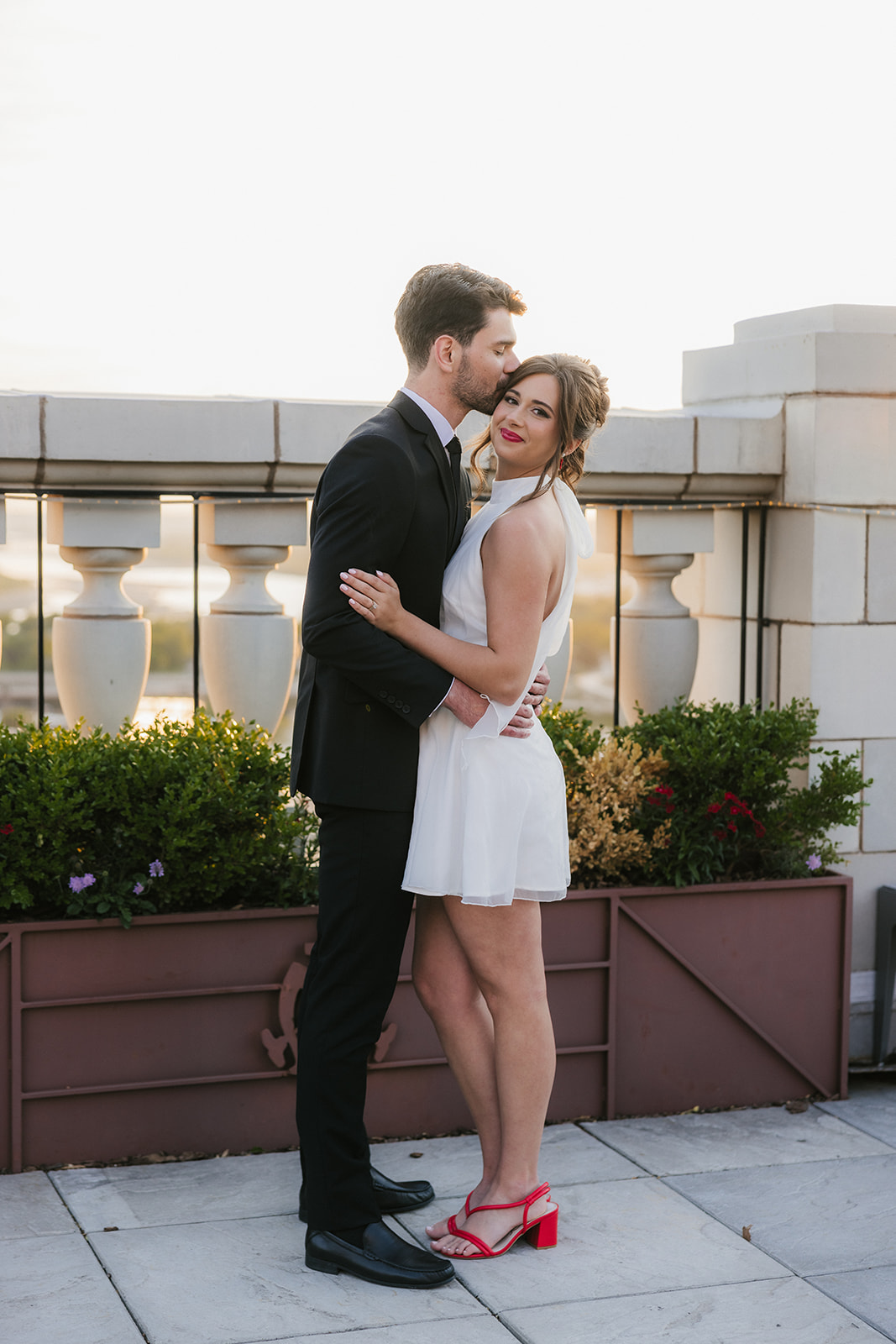 A man in a black suit kisses a woman in a white dress and red heels on the forehead as they stand outdoors on a terrace with decorative railings and planters.