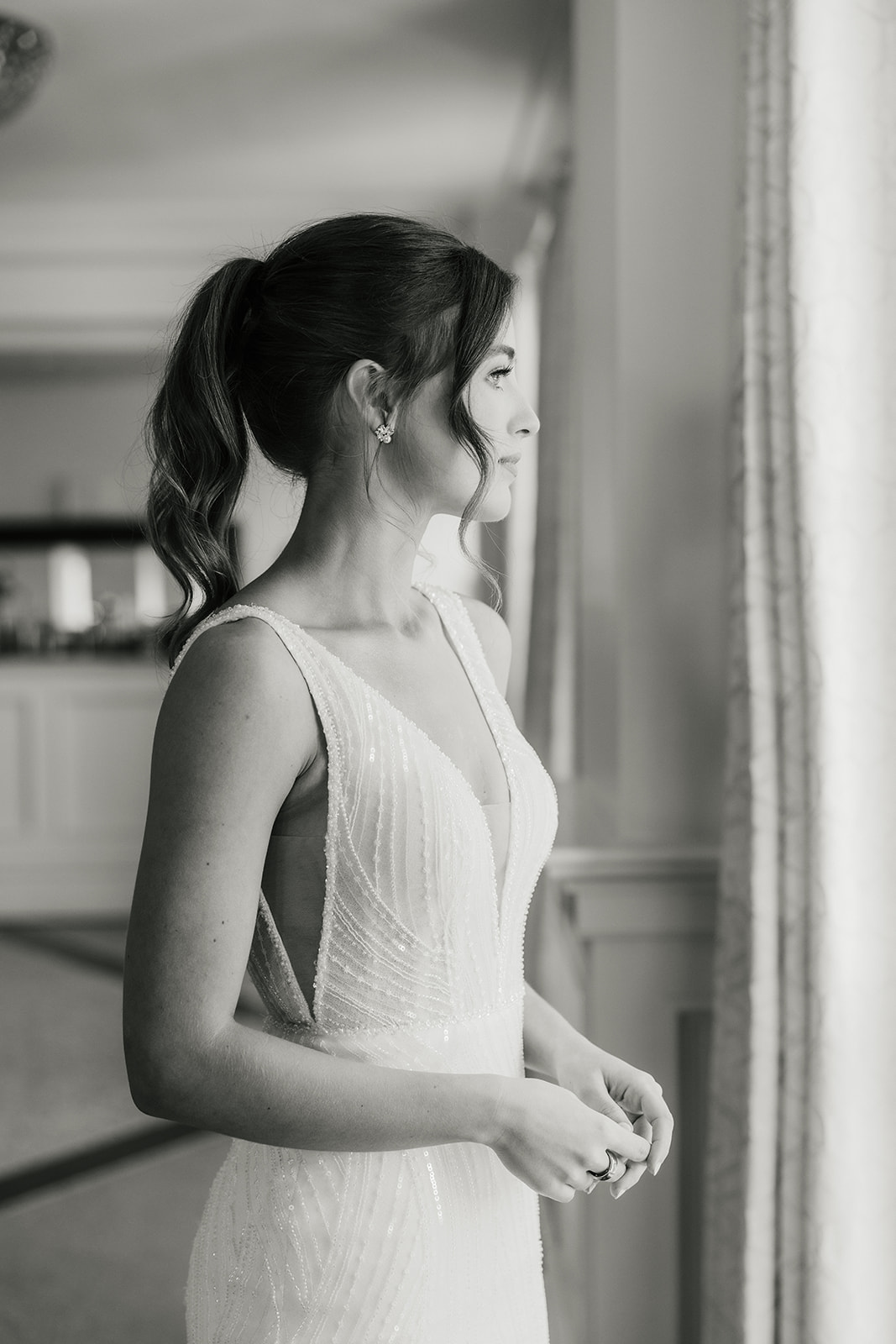 Woman in a white sleeveless dress stands by a window, holding and spraying a perfume bottle, with her hair styled in a ponytail.