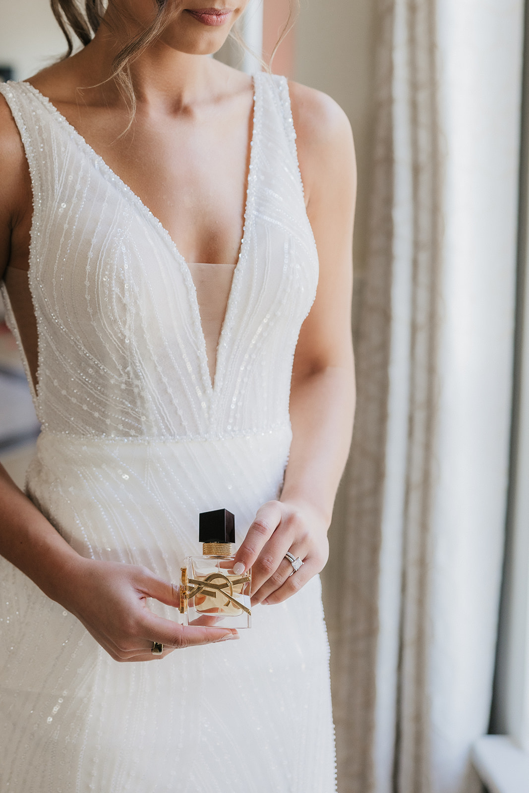 Woman in a white sleeveless dress stands by a window, holding and spraying a perfume bottle, with her hair styled in a ponytail.