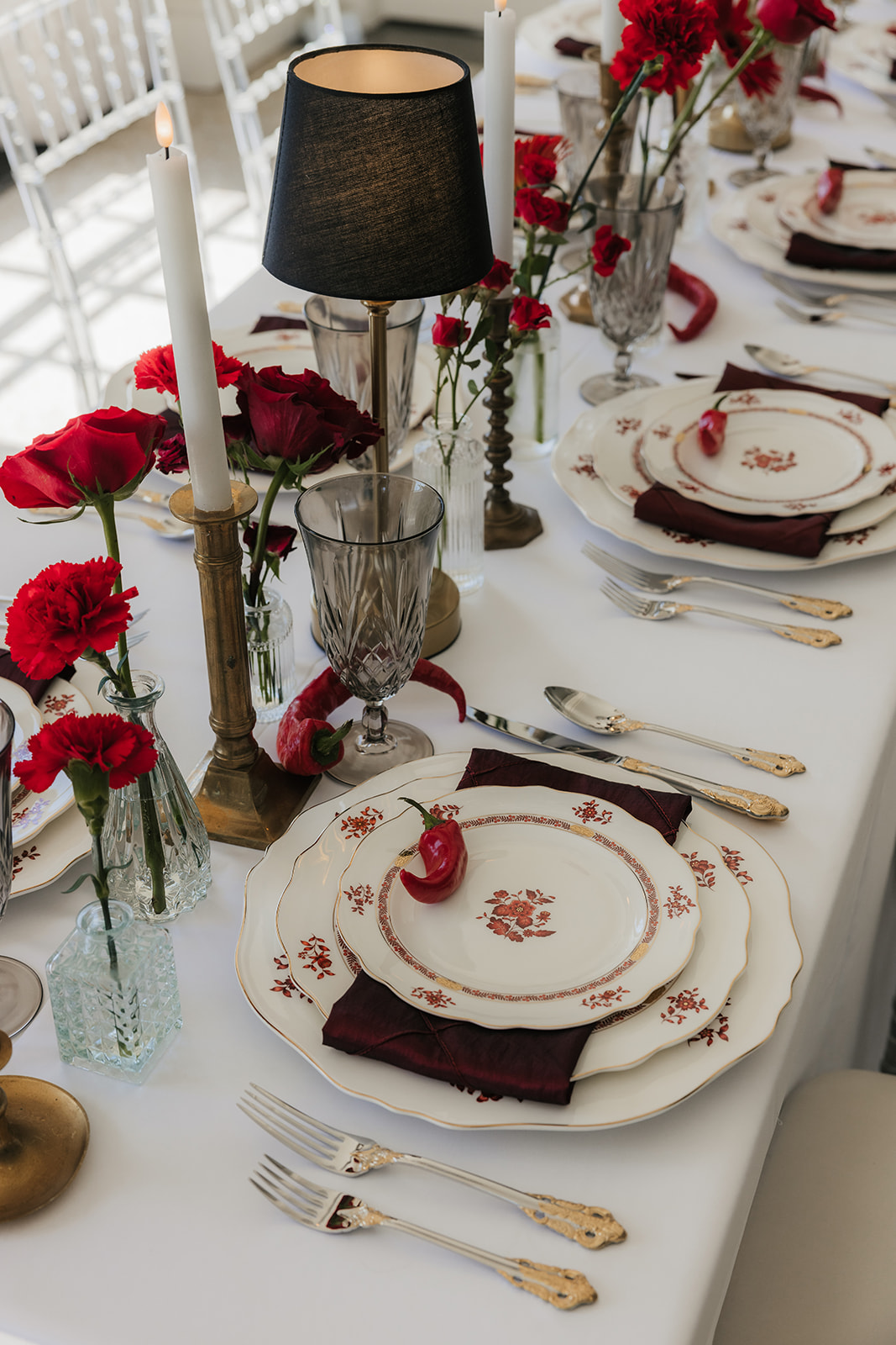 A formal table setting with stacked plates, crystal glassware, gold utensils, red napkin, a red chili pepper on the top plate, and red roses in vases on a white tablecloth for a Mayo Hotel wedding