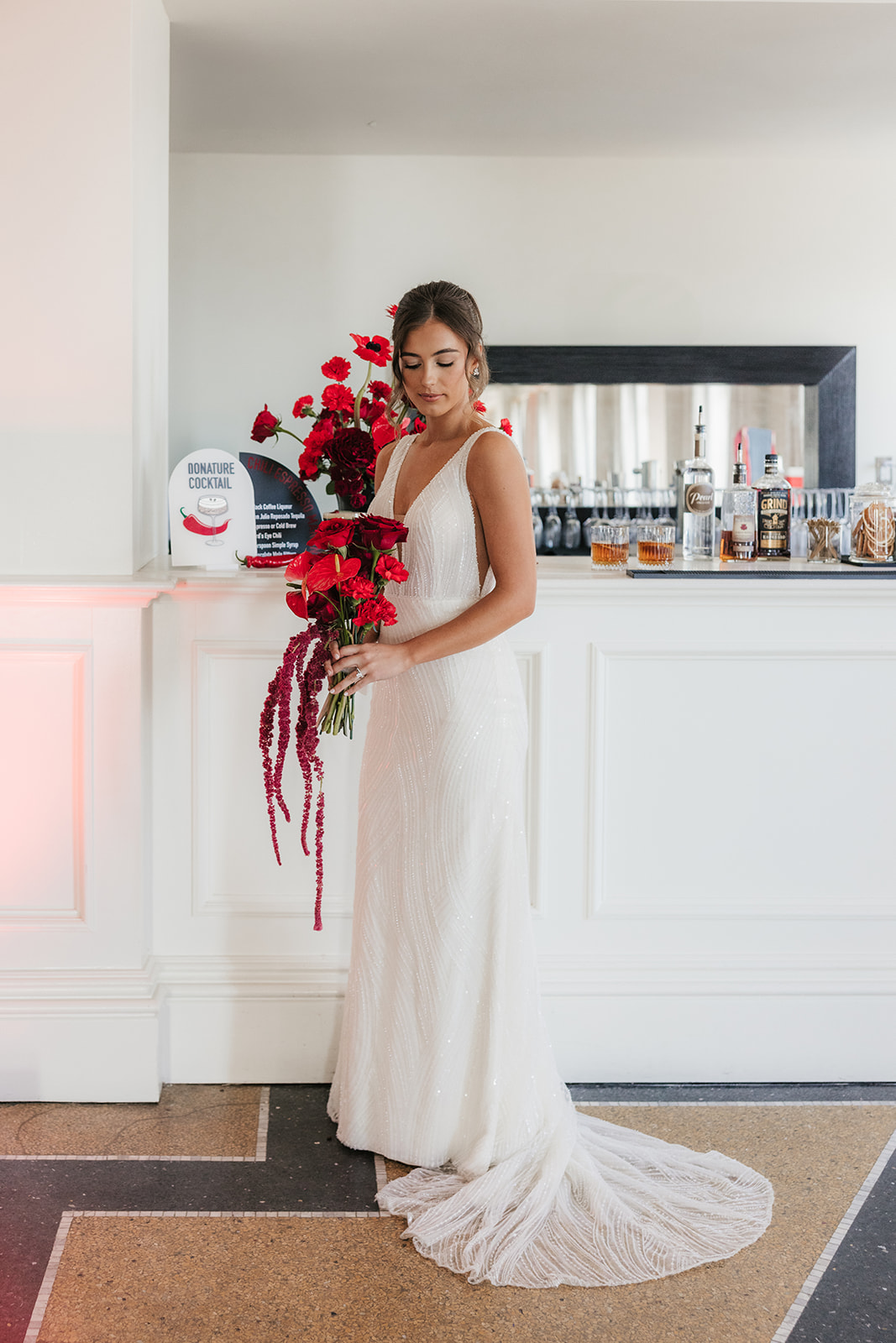 A woman in a white wedding dress stands indoors, holding a large bouquet of red flowers near a bar counter.