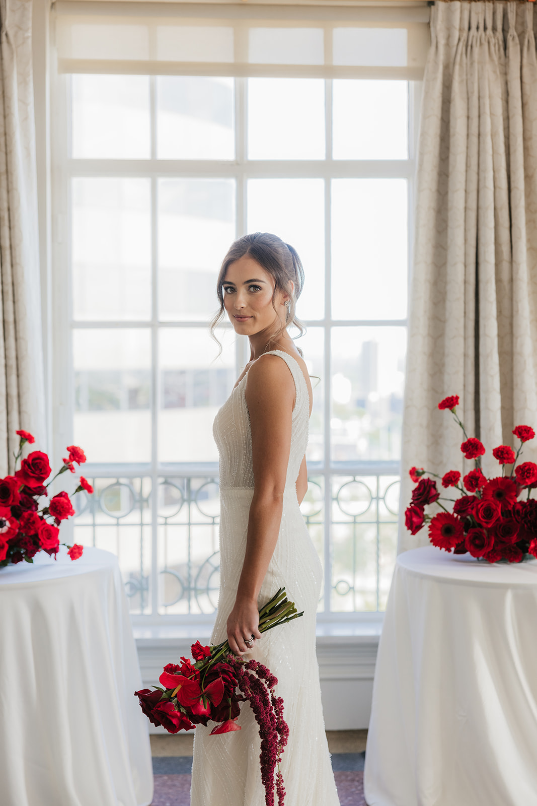 A woman in a white wedding dress stands indoors, holding a large bouquet of red flowers near a bar counter.