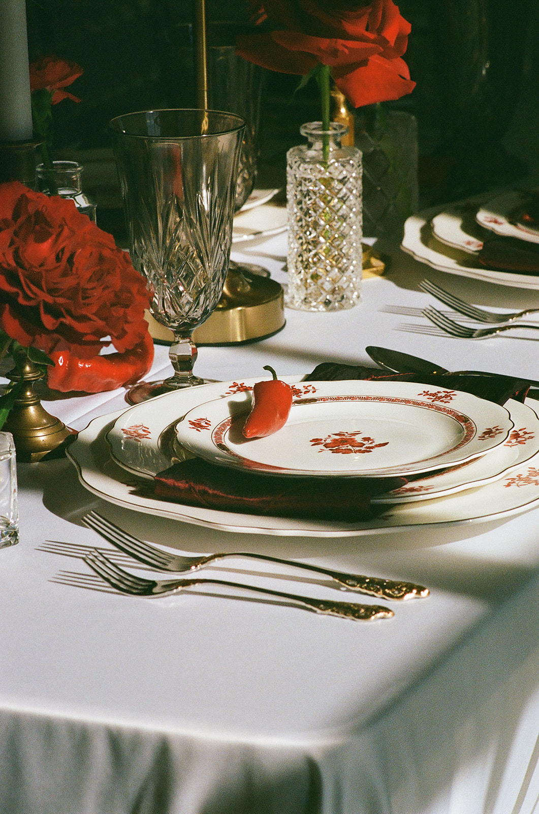 A formal table setting with stacked plates, crystal glassware, gold utensils, red napkin, a red chili pepper on the top plate, and red roses in vases on a white tablecloth for a Mayo Hotel wedding