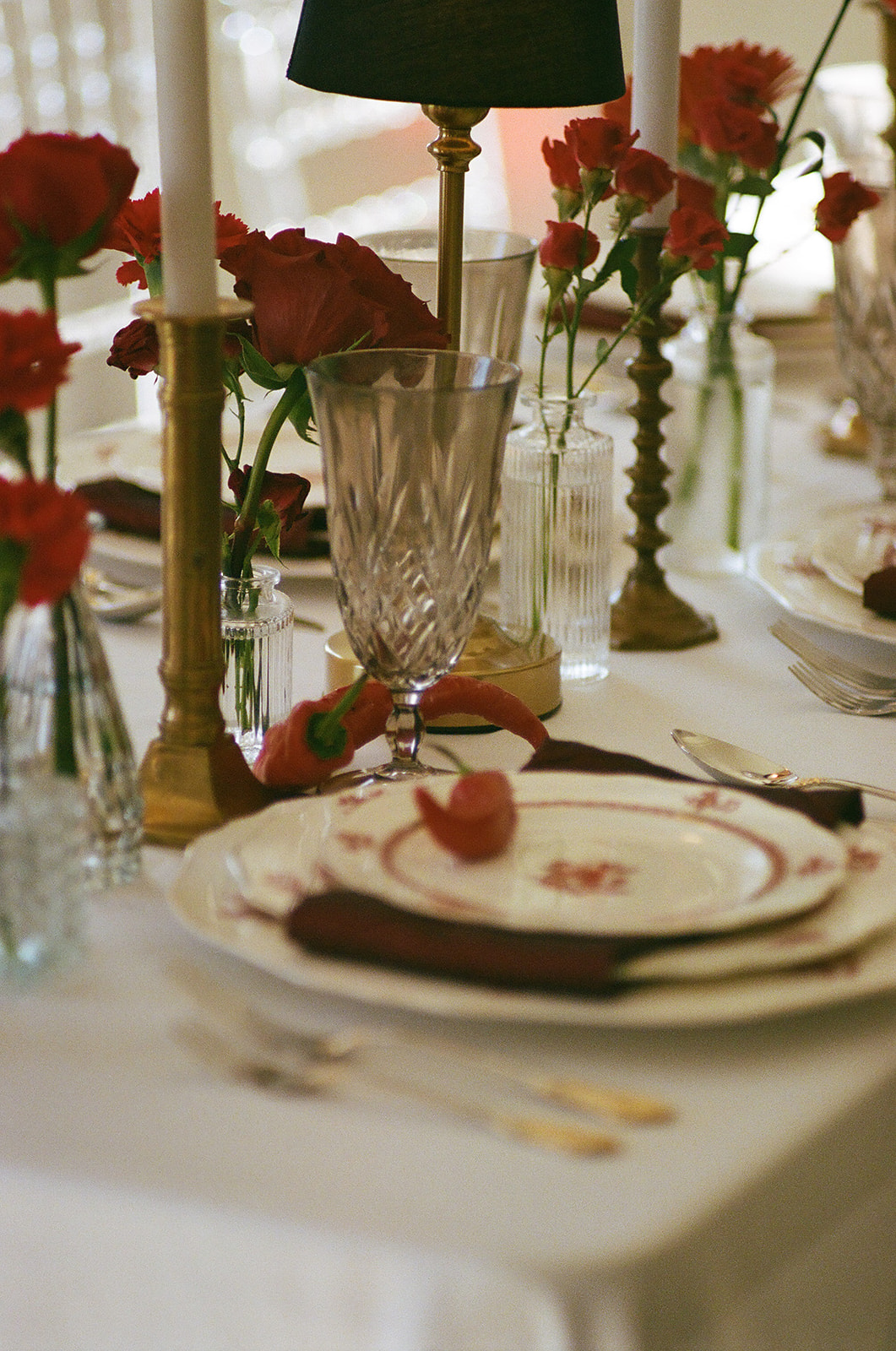 A formal table setting with stacked plates, crystal glassware, gold utensils, red napkin, a red chili pepper on the top plate, and red roses in vases on a white tablecloth for a Mayo Hotel wedding