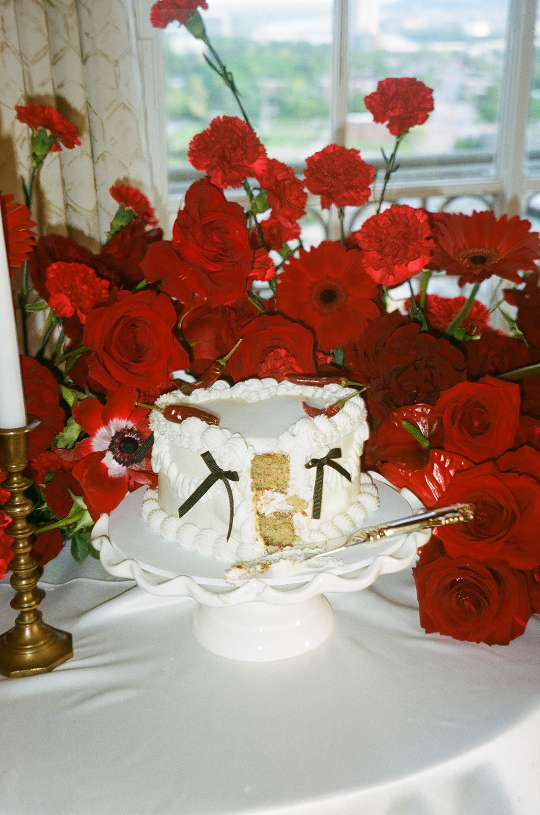 A white frosted cake with a slice cut out is displayed on a cake stand, surrounded by large red flowers and a lit candle on a white tablecloth.