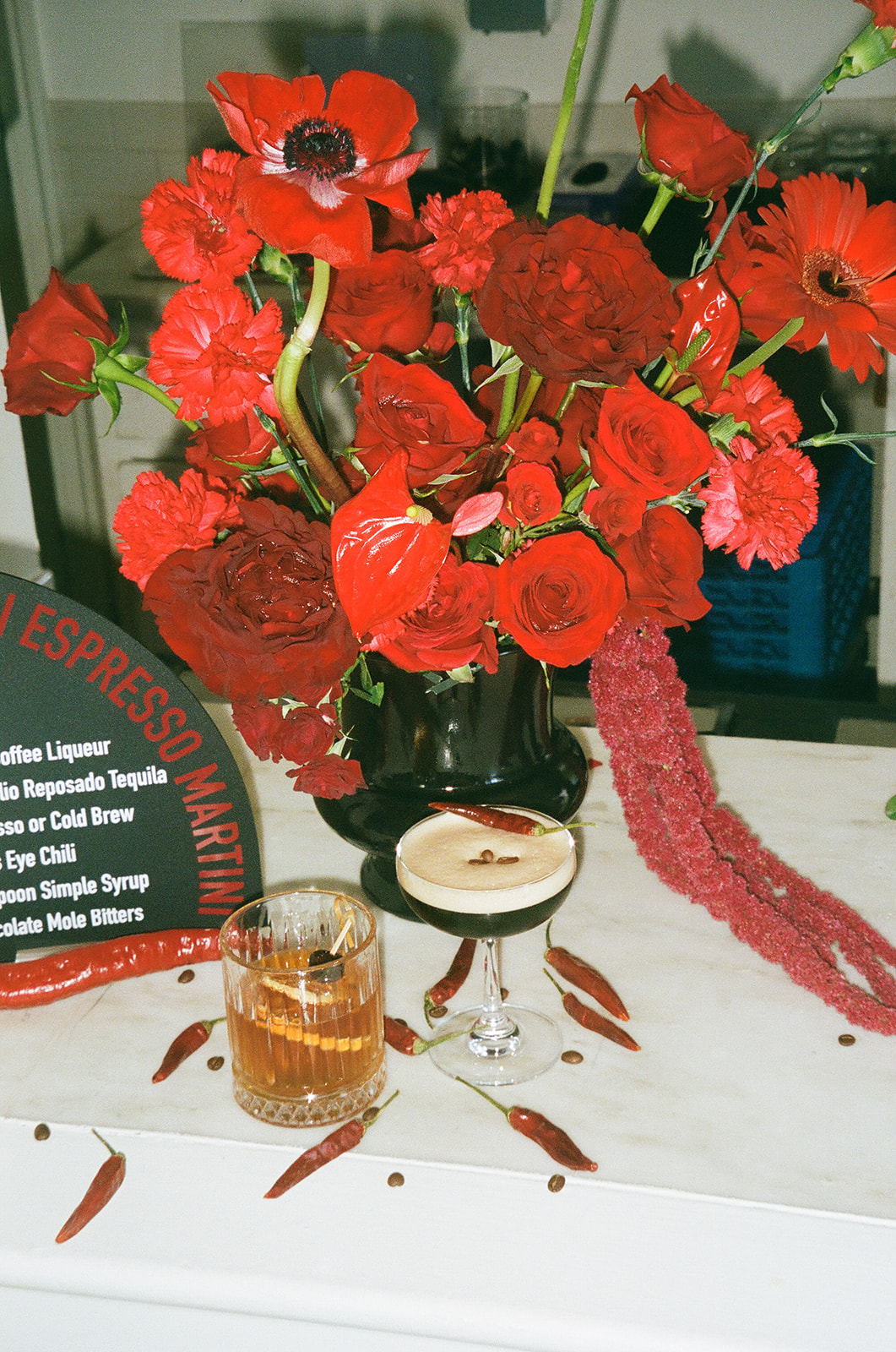 A white frosted cake with a slice cut out is displayed on a cake stand, surrounded by large red flowers and a lit candle on a white tablecloth.