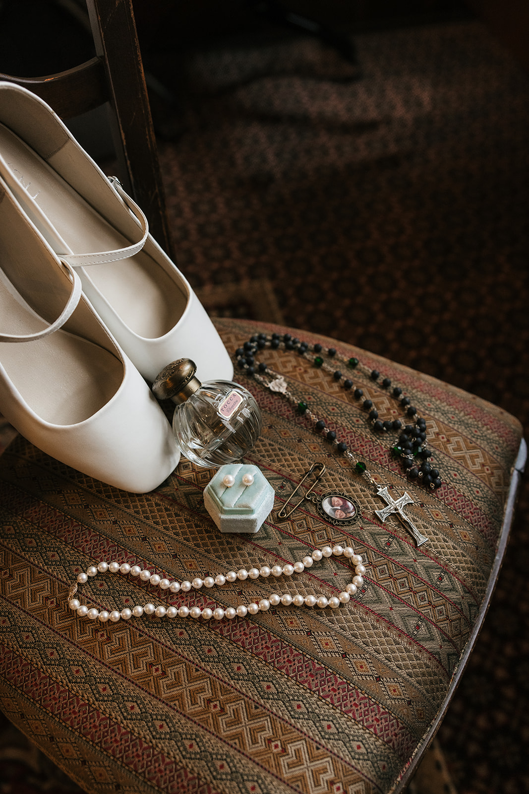 White shoes, a pearl necklace, a rosary, a ring in a blue box, a small perfume bottle, and a pin arranged on an upholstered chair.