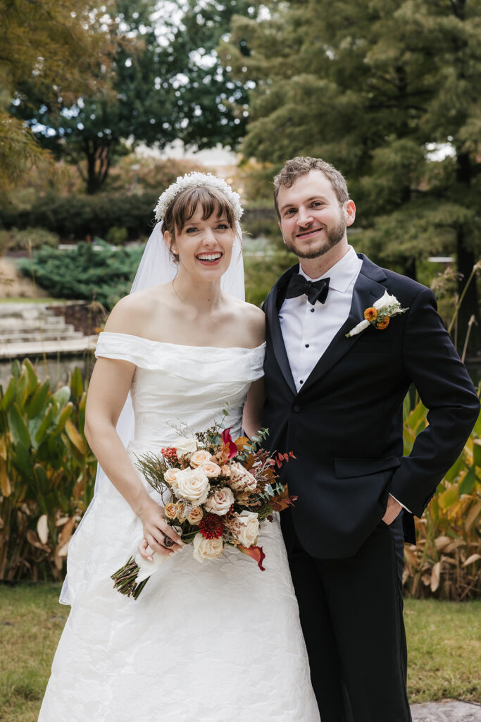 A bride in a white dress and a groom in a black tuxedo pose together outdoors, with greenery and flowers in the background. The bride holds a bouquet captured by a catholic wedding photographer
