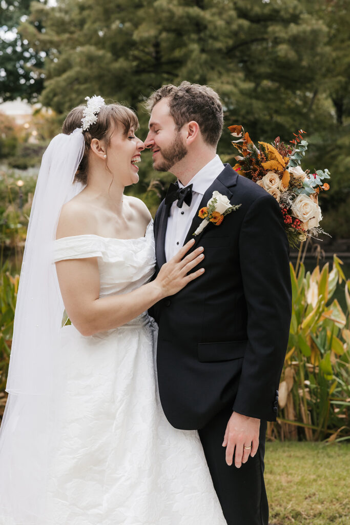A bride in a white dress and a groom in a black tuxedo pose together outdoors, with greenery and flowers in the background. The bride holds a bouquet captured by a catholic wedding photographer