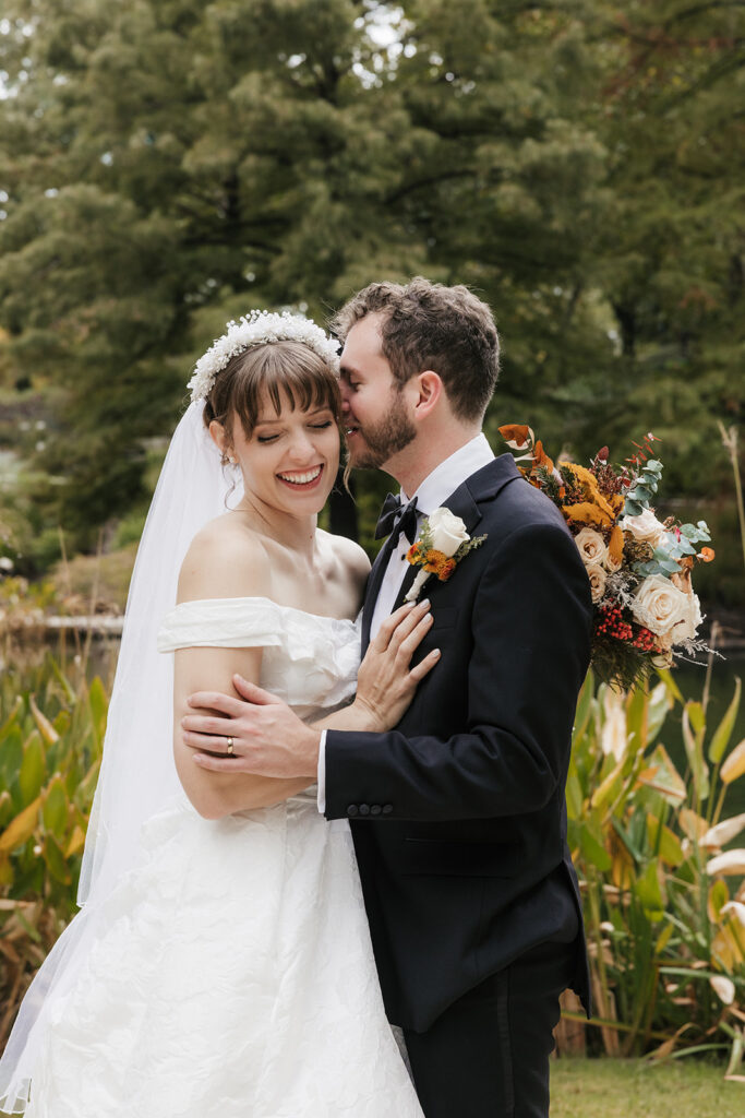 A bride in a white dress and a groom in a black tuxedo pose together outdoors, with greenery and flowers in the background. The bride holds a bouquet captured by a catholic wedding photographer