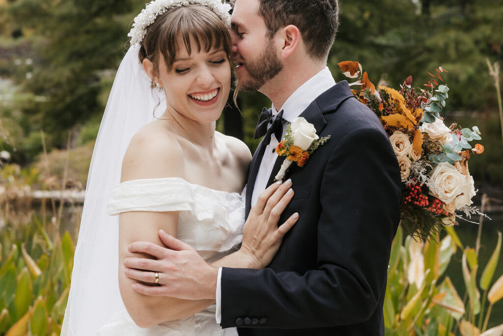 A bride in a white dress and a groom in a black tuxedo pose together outdoors, with greenery and flowers in the background. The bride holds a bouquet captured by a catholic wedding photographer