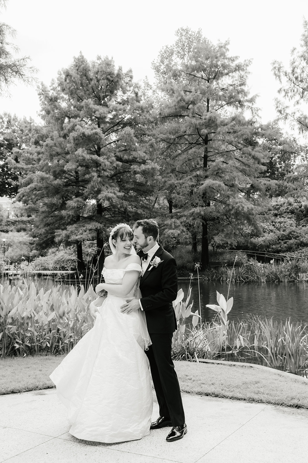 A bride in a white dress and a groom in a black tuxedo pose together outdoors, with greenery and flowers in the background. The bride holds a bouquet captured by a catholic wedding photographer
