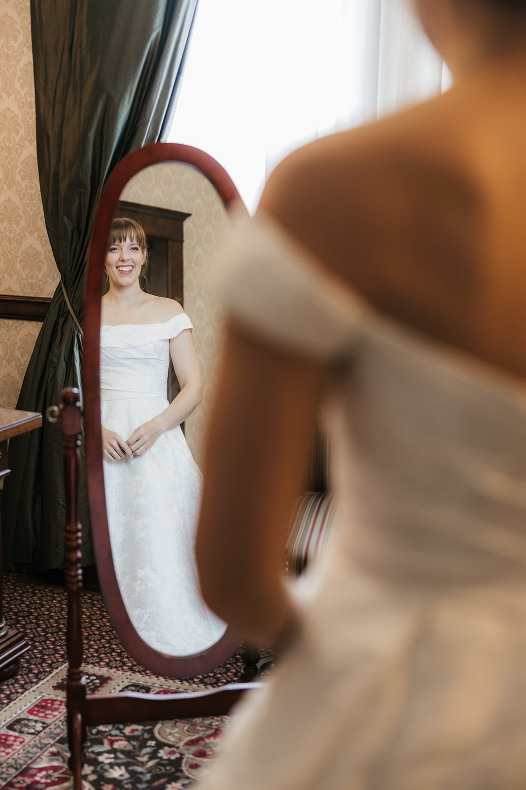 A woman in a white off-the-shoulder dress looks at her reflection in an oval mirror in a carpeted room with patterned wallpaper and drapes captured by catholic wedding photographer