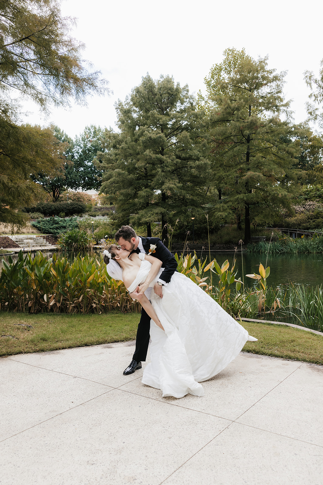 A bride in a white dress and a groom in a black tuxedo pose together outdoors, with greenery and flowers in the background. The bride holds a bouquet captured by a catholic wedding photographer
