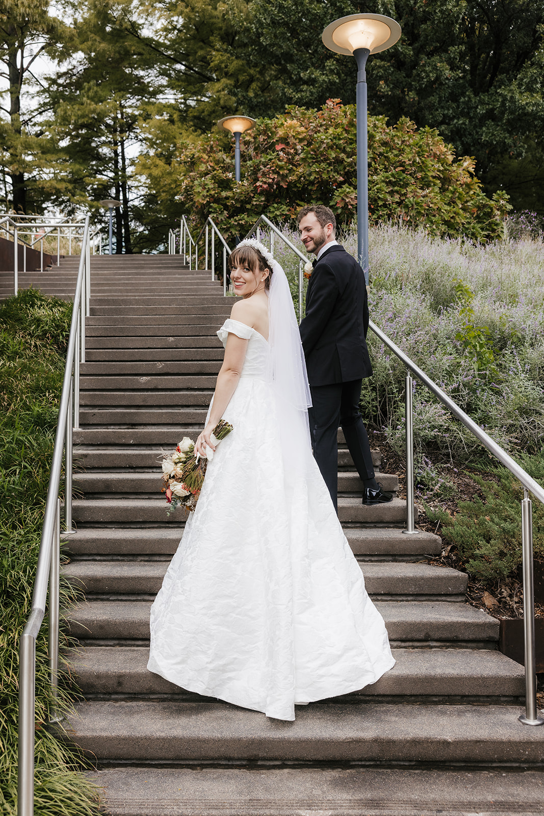 A bride in a white dress and a groom in a black tuxedo pose together outdoors, with greenery and flowers in the background. The bride holds a bouquet captured by a catholic wedding photographer