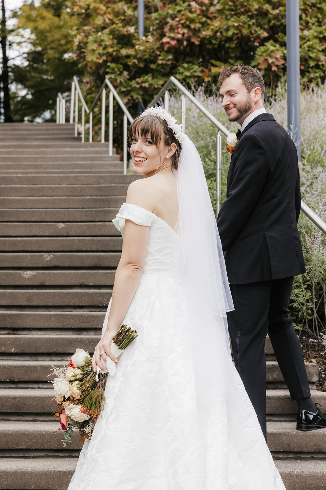 A bride in a white dress and a groom in a black tuxedo pose together outdoors, with greenery and flowers in the background. The bride holds a bouquet captured by a catholic wedding photographer