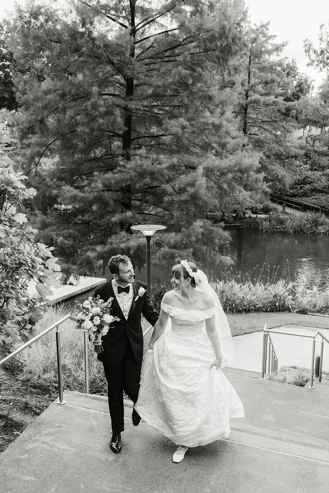 A bride in a white dress and a groom in a black tuxedo pose together outdoors, with greenery and flowers in the background. The bride holds a bouquet captured by a catholic wedding photographer