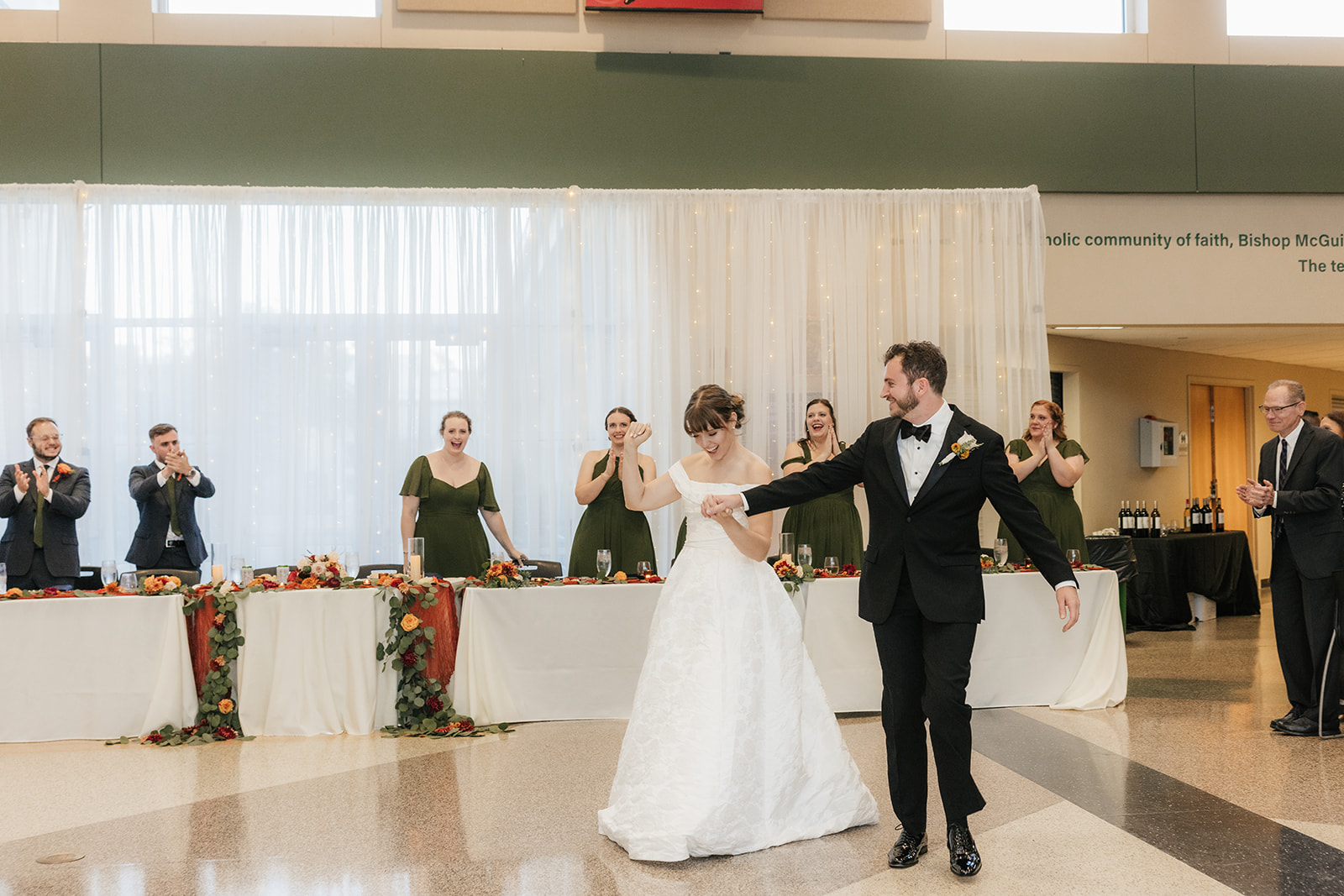 A bride and groom dance together in front of a decorated table, while their wedding party stands and applauds in the background.