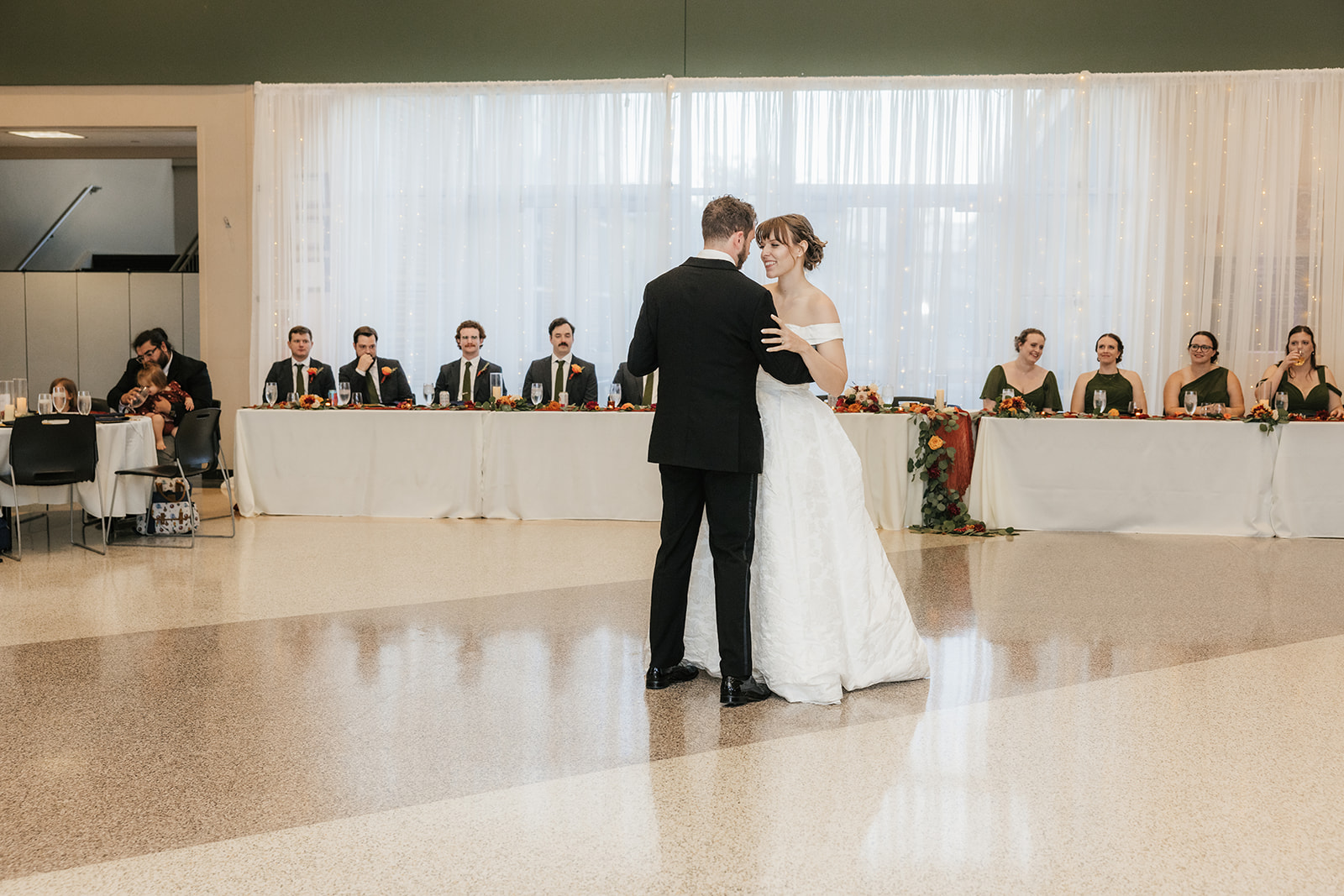 A bride and groom dance together in front of a decorated table, while their wedding party stands and applauds in the background.