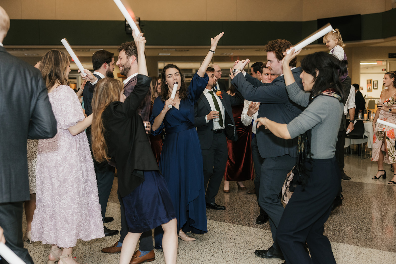 A group of people in formal attire dance indoors, holding light sticks and raising their arms, with others mingling in the background.