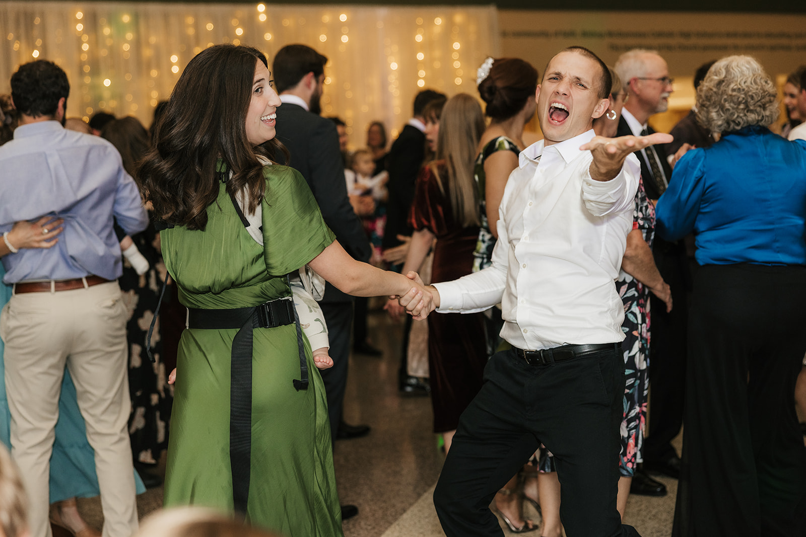A group of people in formal attire dance indoors, holding light sticks and raising their arms, with others mingling in the background.