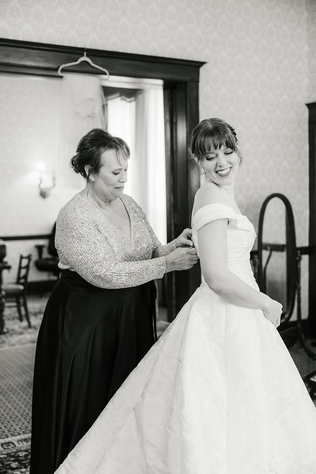 A woman helps another woman adjust the back of her off-the-shoulder wedding dress in a room with classic decor captured by a catholic wedding photographer