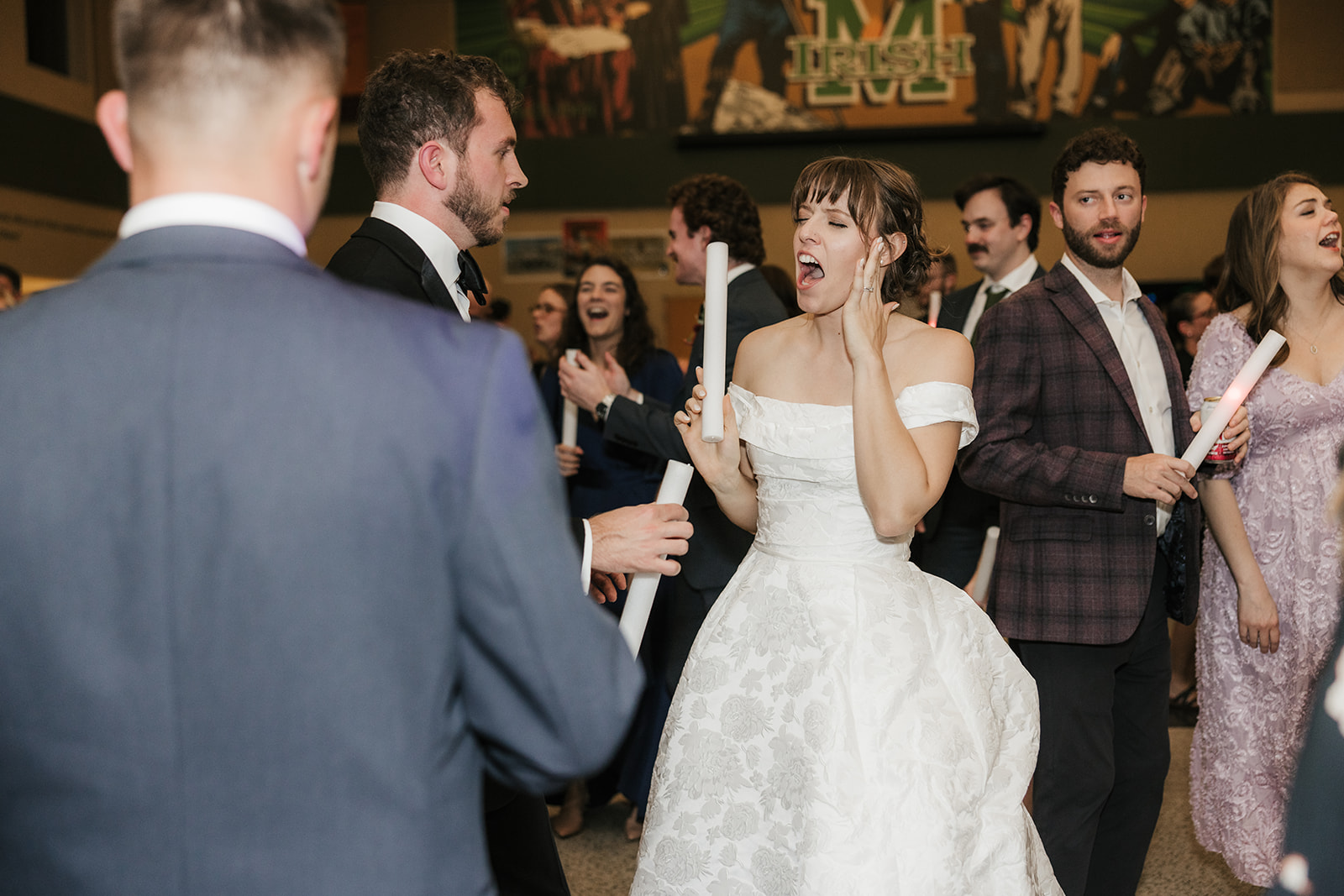 A group of people in formal attire dance indoors, holding light sticks and raising their arms, with others mingling in the background.