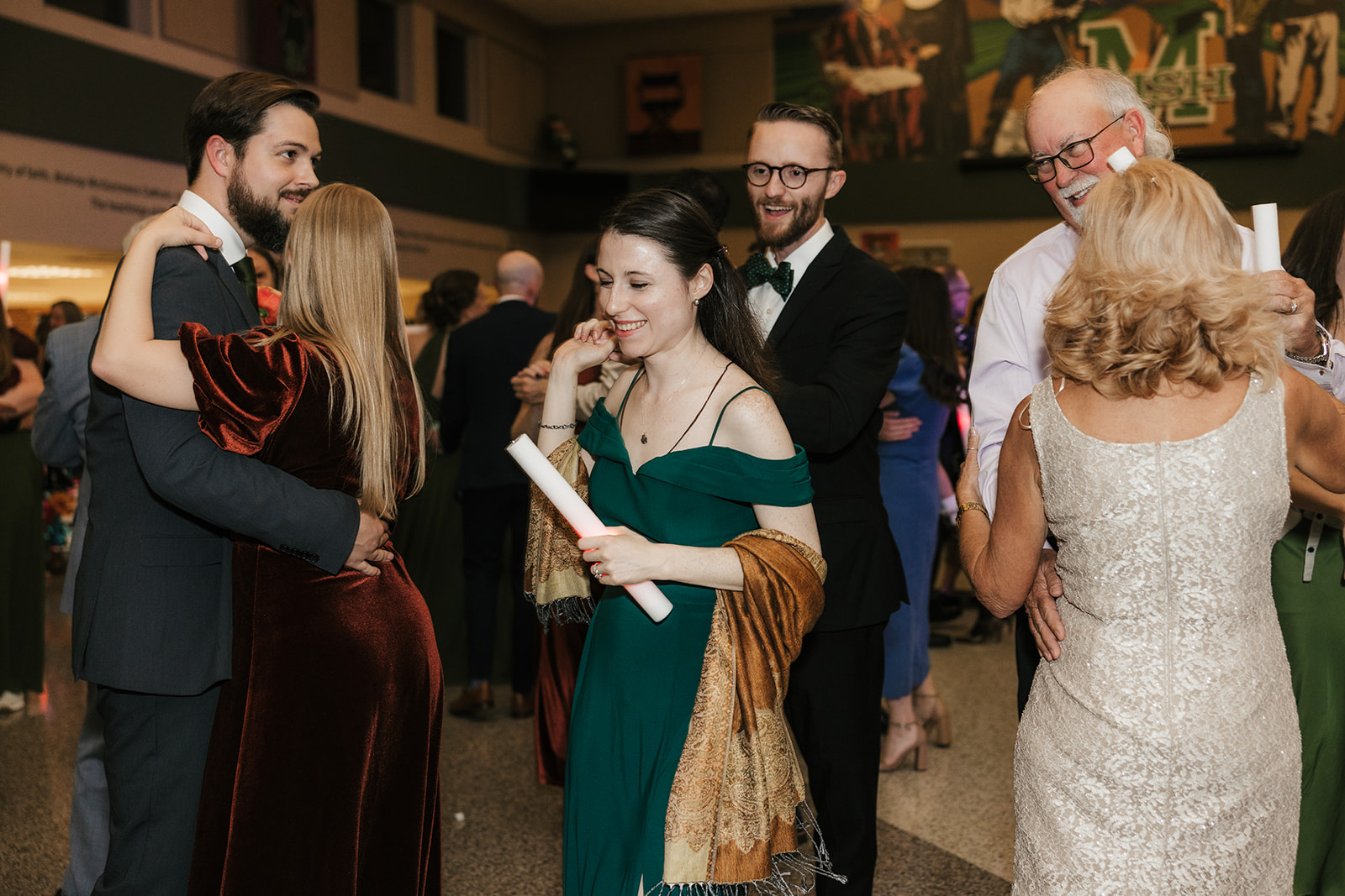 A group of people in formal attire dance indoors, holding light sticks and raising their arms, with others mingling in the background.