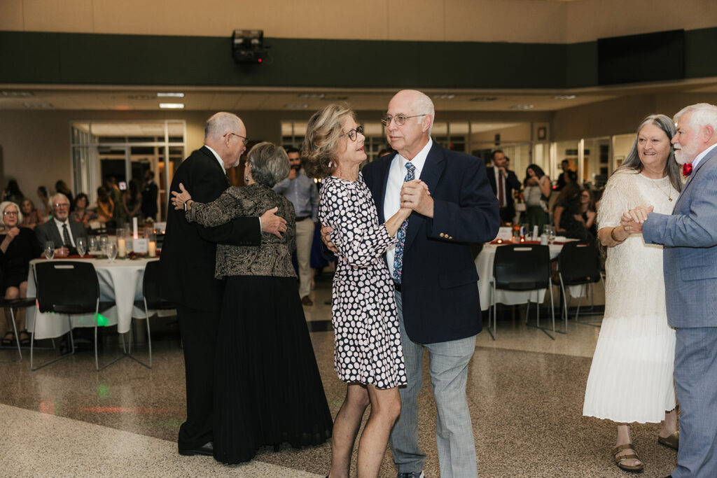 A group of people in formal attire dance indoors, holding light sticks and raising their arms, with others mingling in the background.
