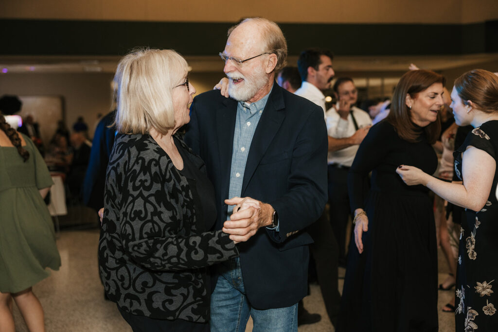 A bride in a white gown dances with guests in formal attire at a wedding reception, while other people socialize in the background.
