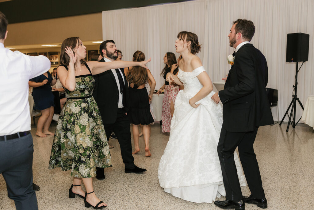 A bride in a white gown dances with guests in formal attire at a wedding reception, while other people socialize in the background.