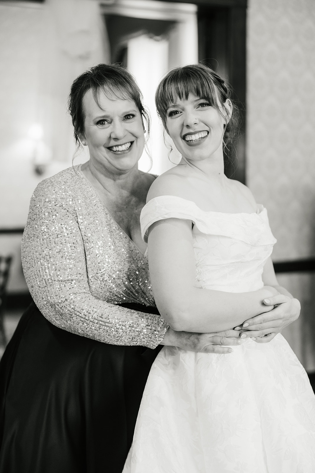 A woman helps another woman adjust the back of her off-the-shoulder wedding dress in a room with classic decor captured by a catholic wedding photographer