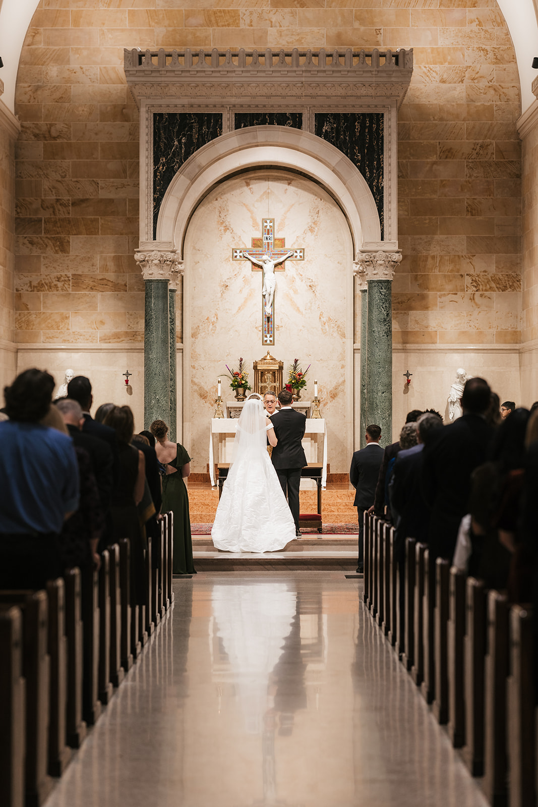 A bride and groom stand at the altar in a church during their wedding ceremony, with guests seated on both sides captured by a catholic wedding photographer