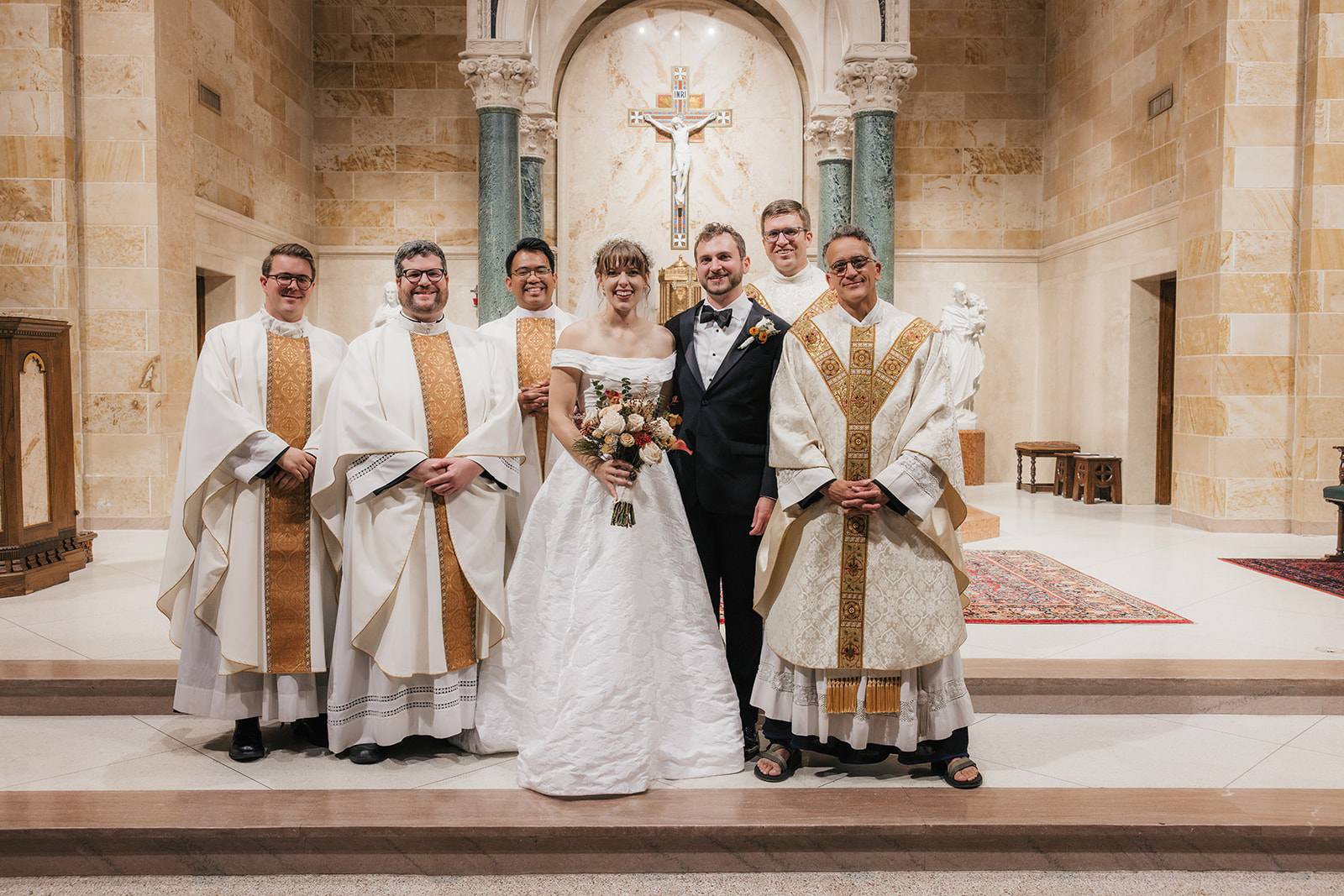 A bride and groom stand together in a church with five clergy members, all dressed in formal attire, posing for a wedding photo.