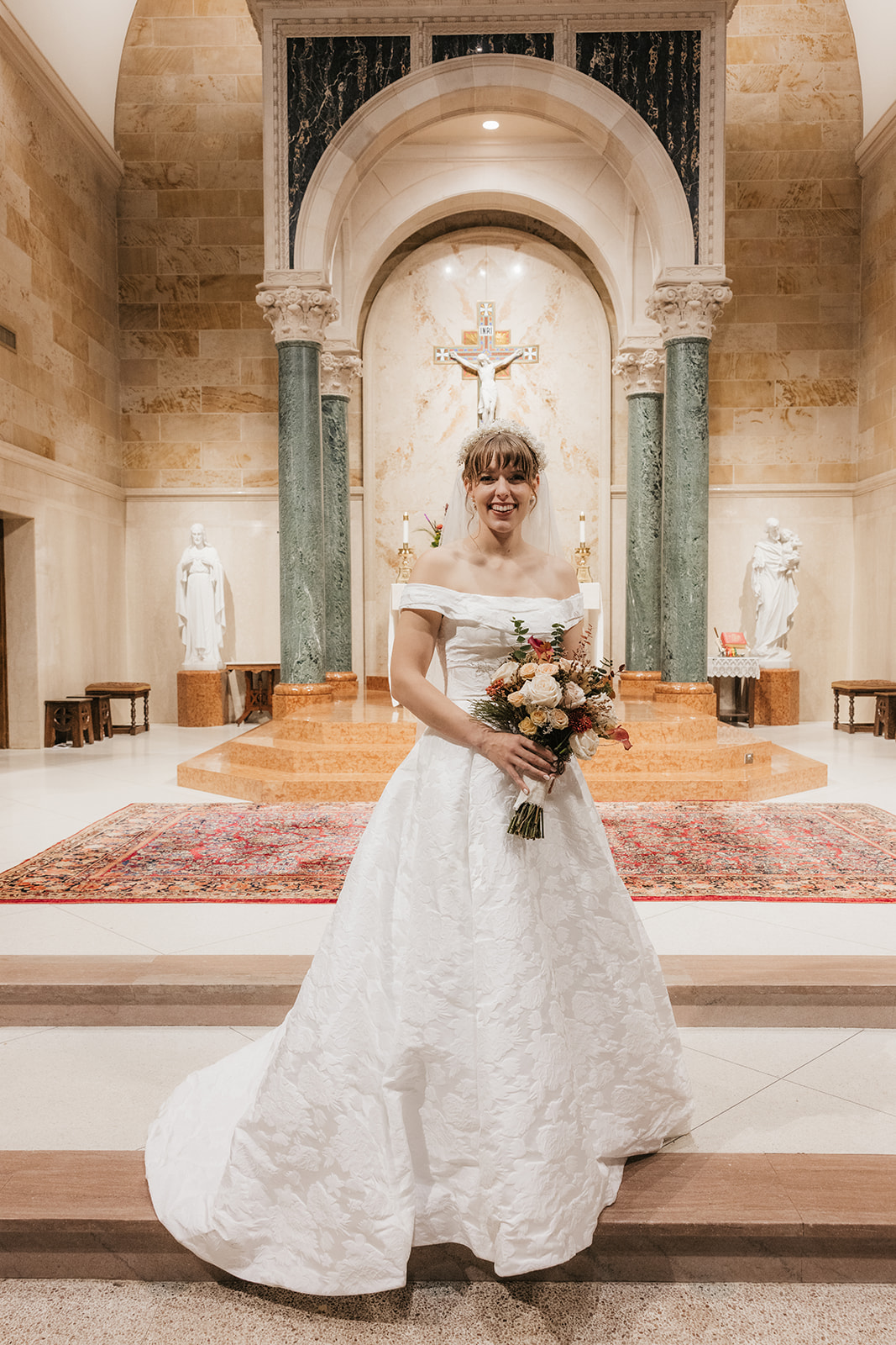 A bride in a white wedding dress holding a bouquet stands in a church sanctuary with statues and an ornate altar in the background captured by a catholic wedding photographer