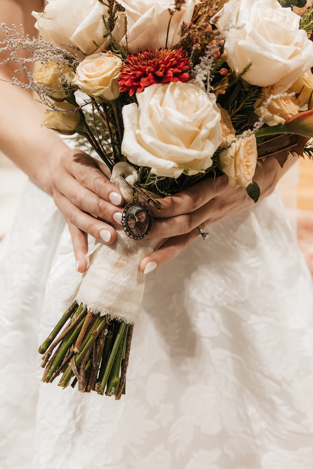 A bride in a white dress holds a bouquet of cream and yellow roses, accented with red flowers and greenery, tied with lace ribbon.