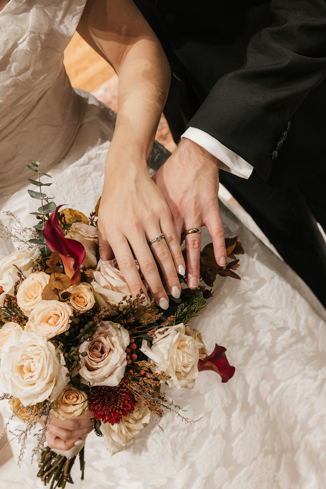 A bride in a white dress holds a bouquet of cream and yellow roses, accented with red flowers and greenery, tied with lace ribbon.