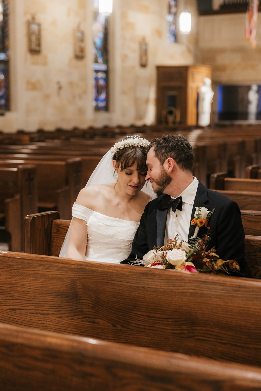 A bride and groom sit closely together in a church pew, touching foreheads and holding a bouquet, with empty pews and stained glass windows in the background captured by a catholic wedding photographer