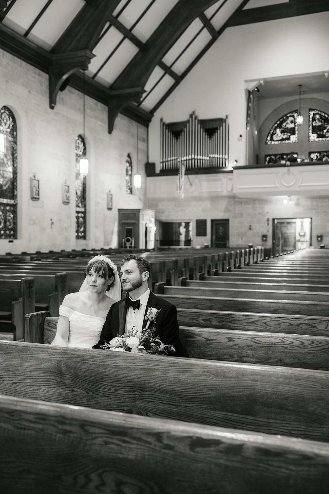 A bride and groom sit closely together in a church pew, touching foreheads and holding a bouquet, with empty pews and stained glass windows in the background captured by a catholic wedding photographer