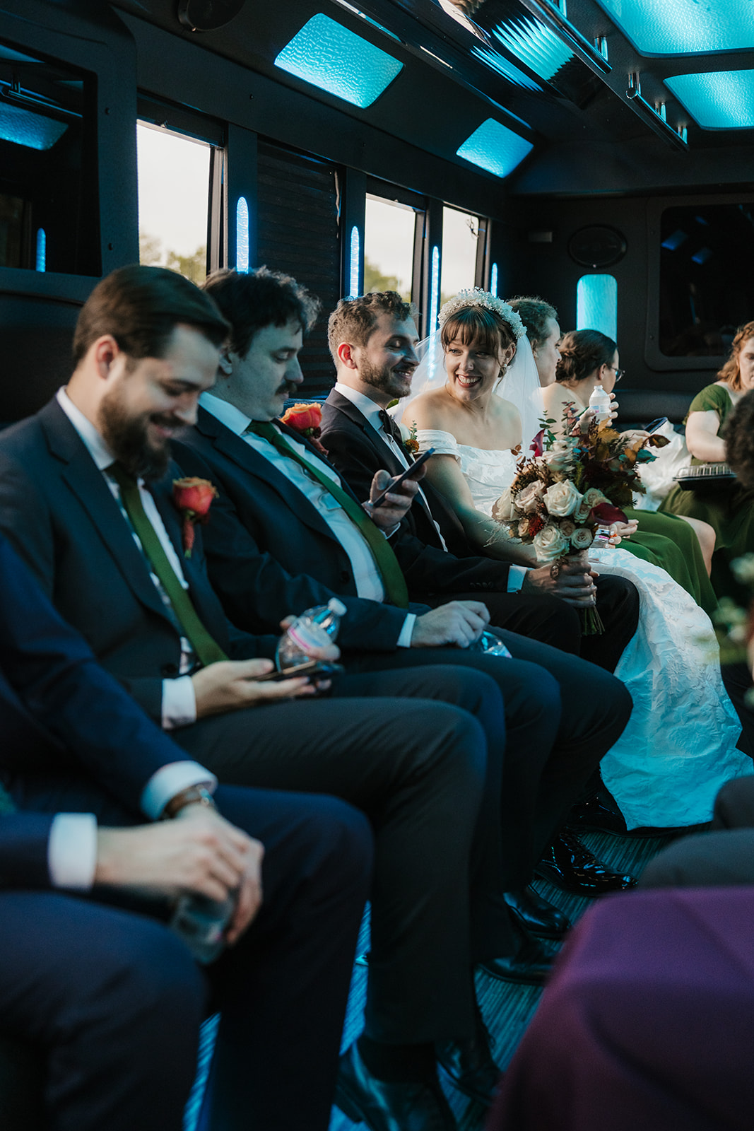 People dressed in formal attire, including a bride and groom, sit in a row inside a vehicle, some holding phones and bouquets.