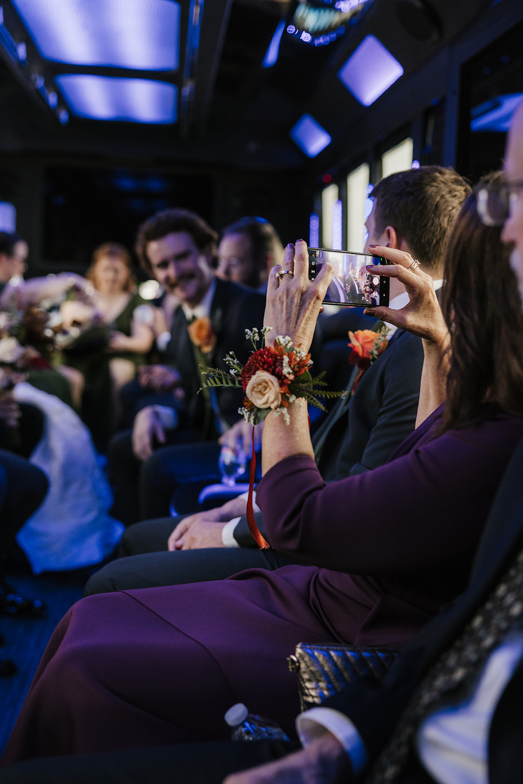 People dressed in formal attire, including a bride and groom, sit in a row inside a vehicle, some holding phones and bouquets.