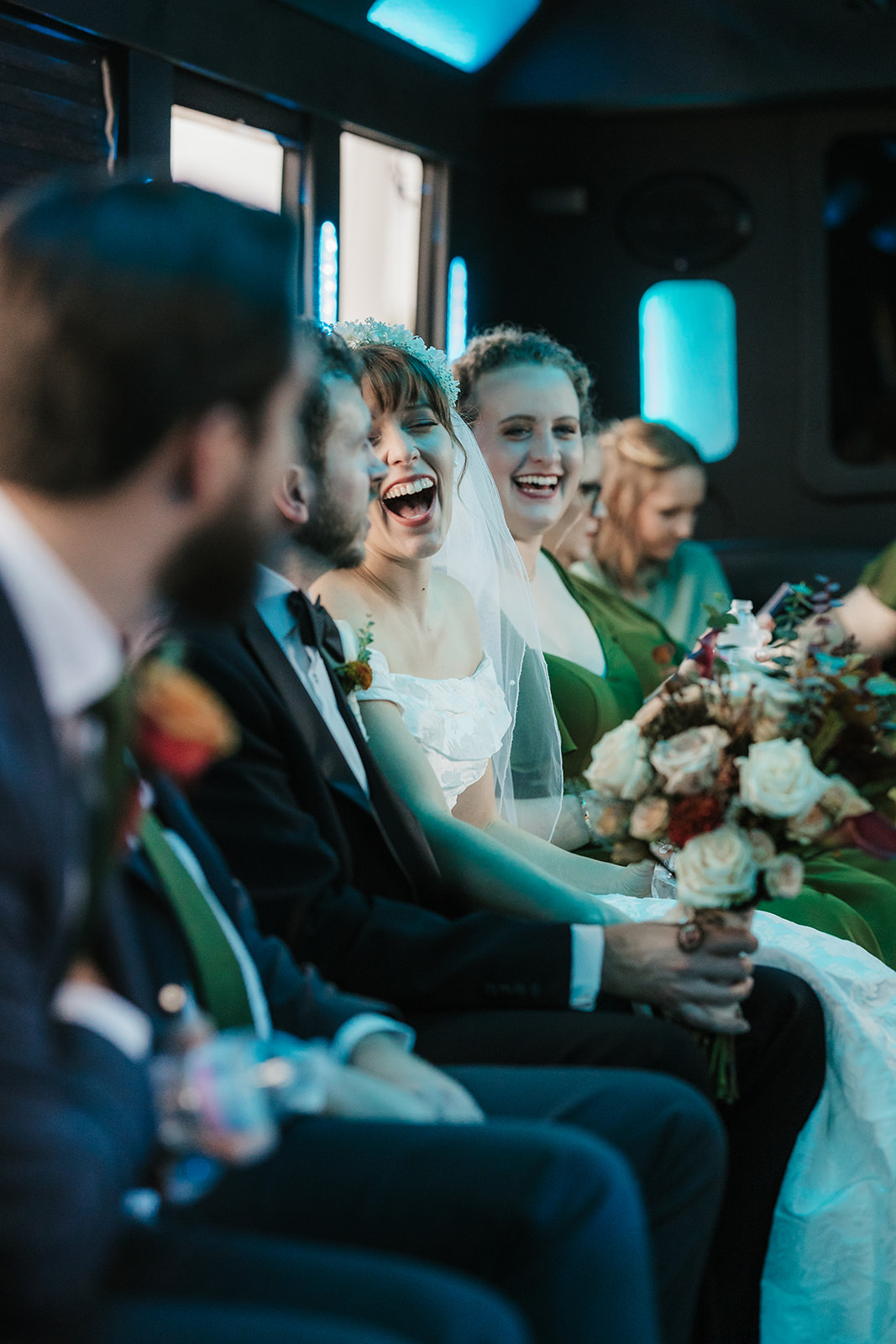 People dressed in formal attire, including a bride and groom, sit in a row inside a vehicle, some holding phones and bouquets.