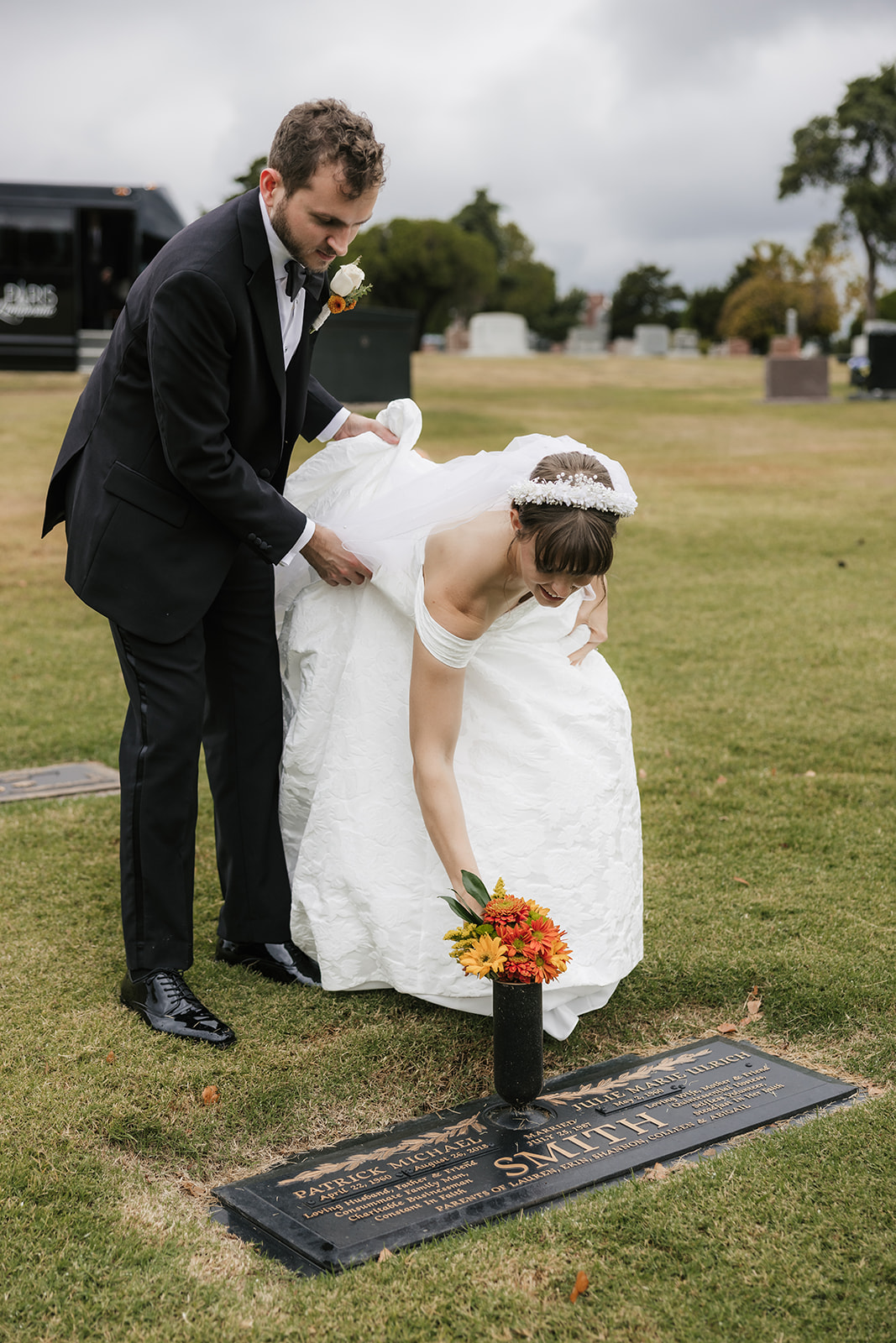 A bride and groom in wedding attire stand together in a grassy cemetery near a grave marker with a bouquet of orange flowers.