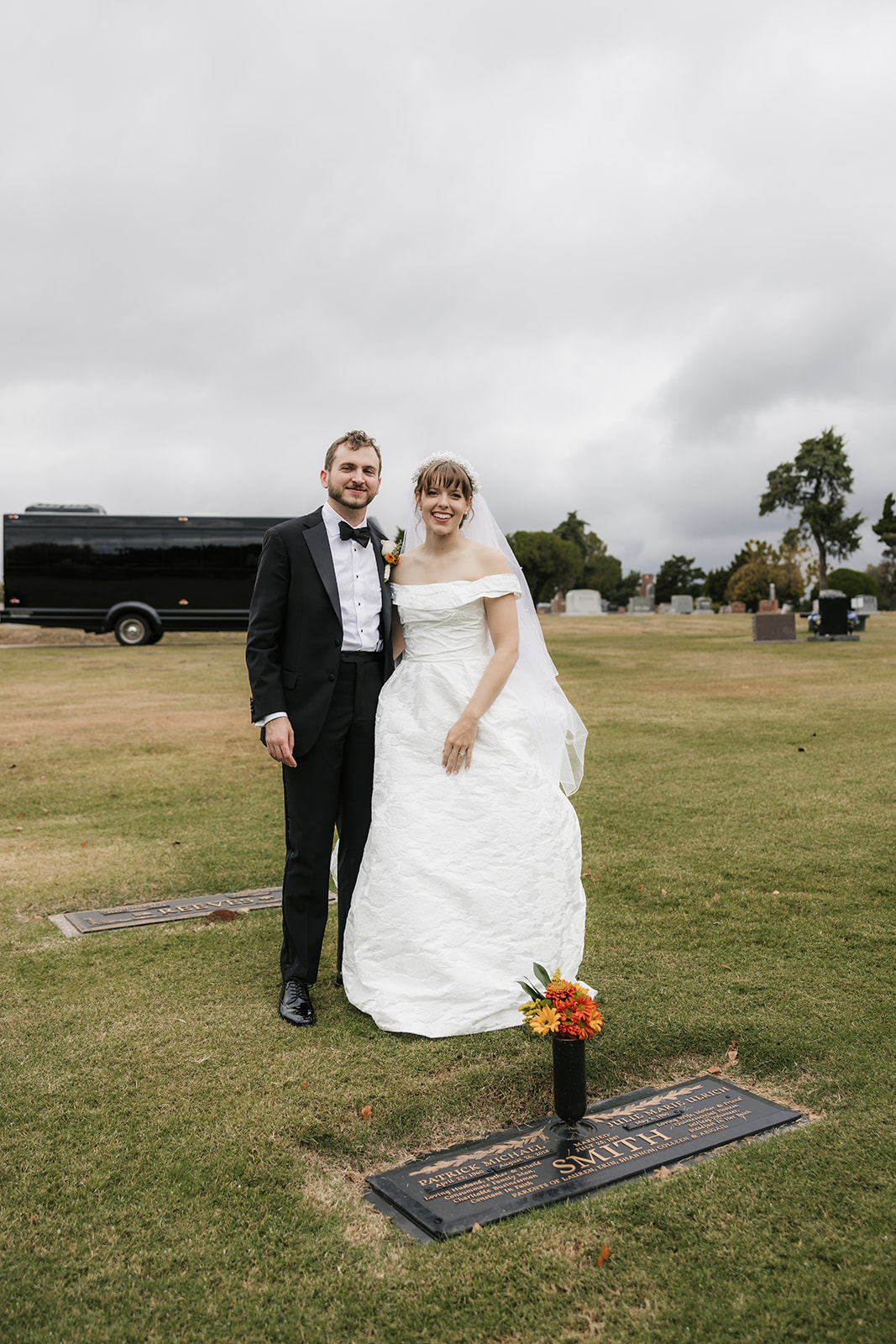A bride and groom in wedding attire stand together in a grassy cemetery near a grave marker with a bouquet of orange flowers.