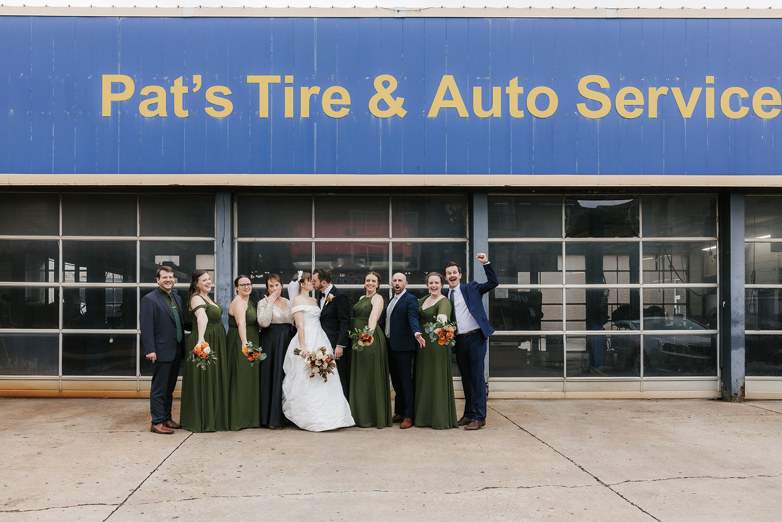 A wedding party poses in front of a Pat's Tire & Auto Service shop, with the bride and groom in the center and bridesmaids and groomsmen on either side.