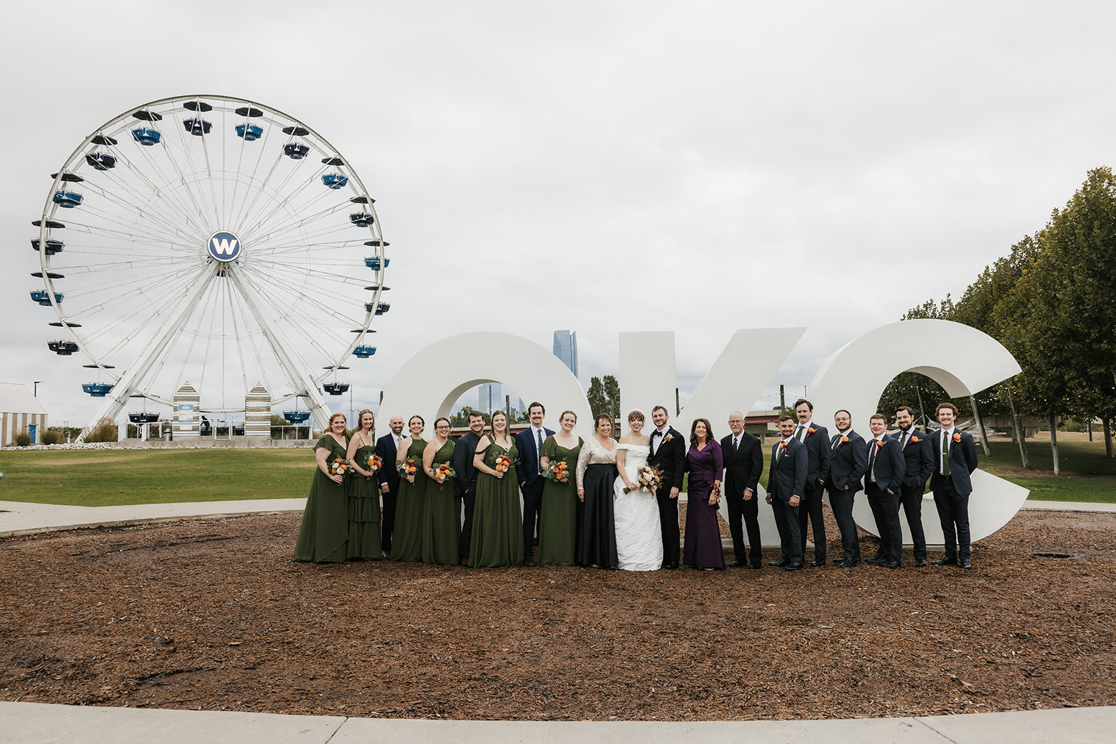 A wedding party poses in front of a large "OKC" sign with a Ferris wheel in the background on a cloudy day.