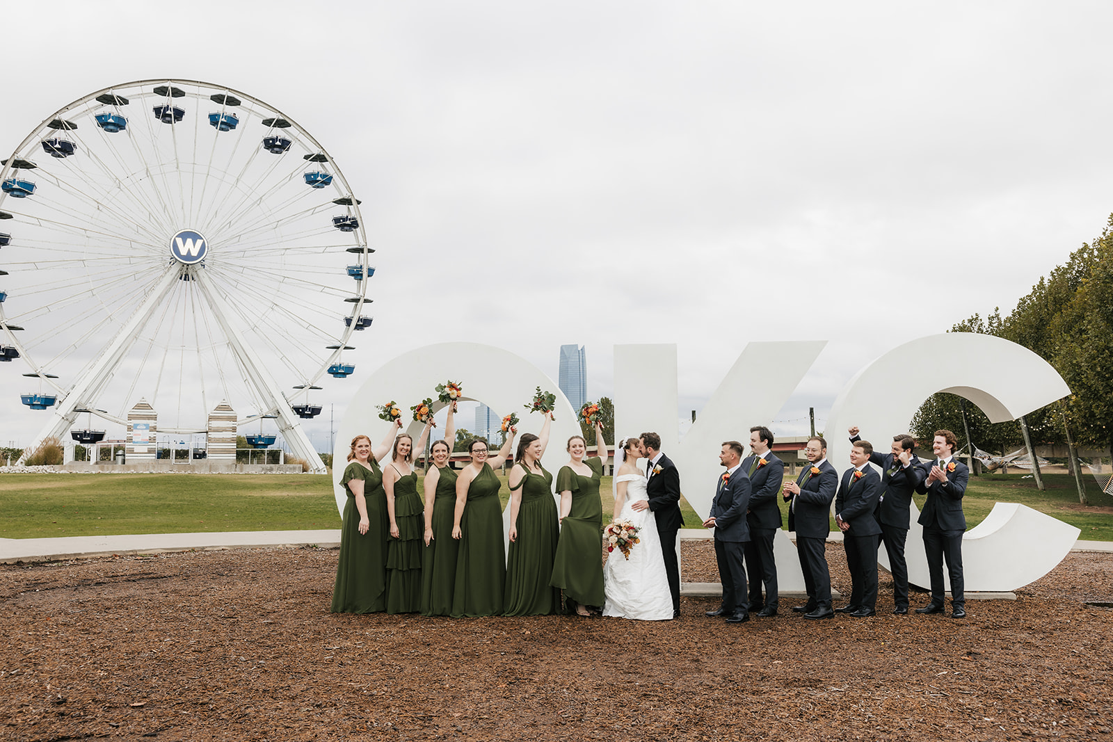 A wedding party poses in front of a large "OKC" sign with a Ferris wheel in the background on a cloudy day.