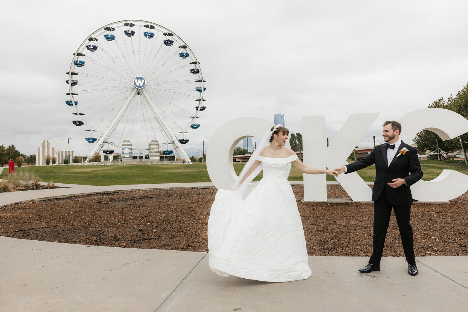 A bride and groom stand in front of large "OKC" letters and a Ferris wheel, holding hands outdoors on a cloudy day.